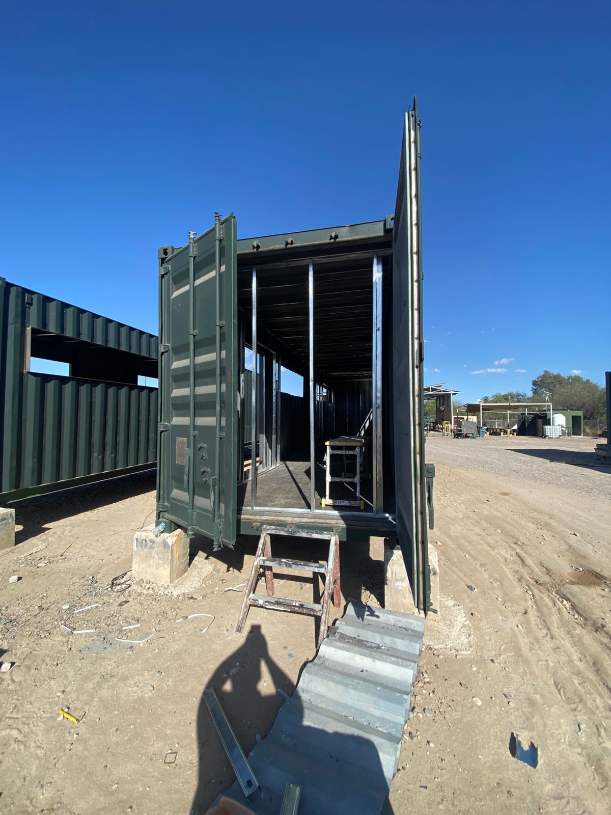 Vista exterior del contenedor verde militar con puertas originales abiertas y rampa metálica de acceso. Se aprecia el corte lateral grande para ventana corrida y la estructura interior ya armada. Al fondo otro contenedor con corte de ventana, sobre la base de concreto numerada 102.