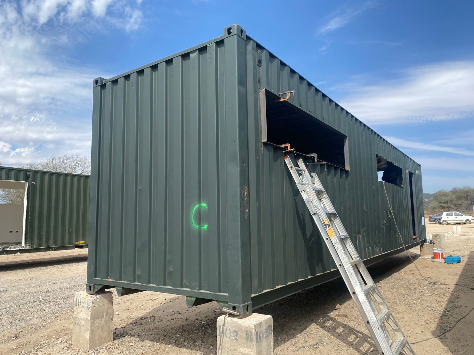 Toma exterior a tres cuartos de un contenedor verde High Cube sobre dados de concreto, con cortes recientes para ventanal y puerta visibles en el costado largo. Marca de spray verde indica nomenclatura de obra.
