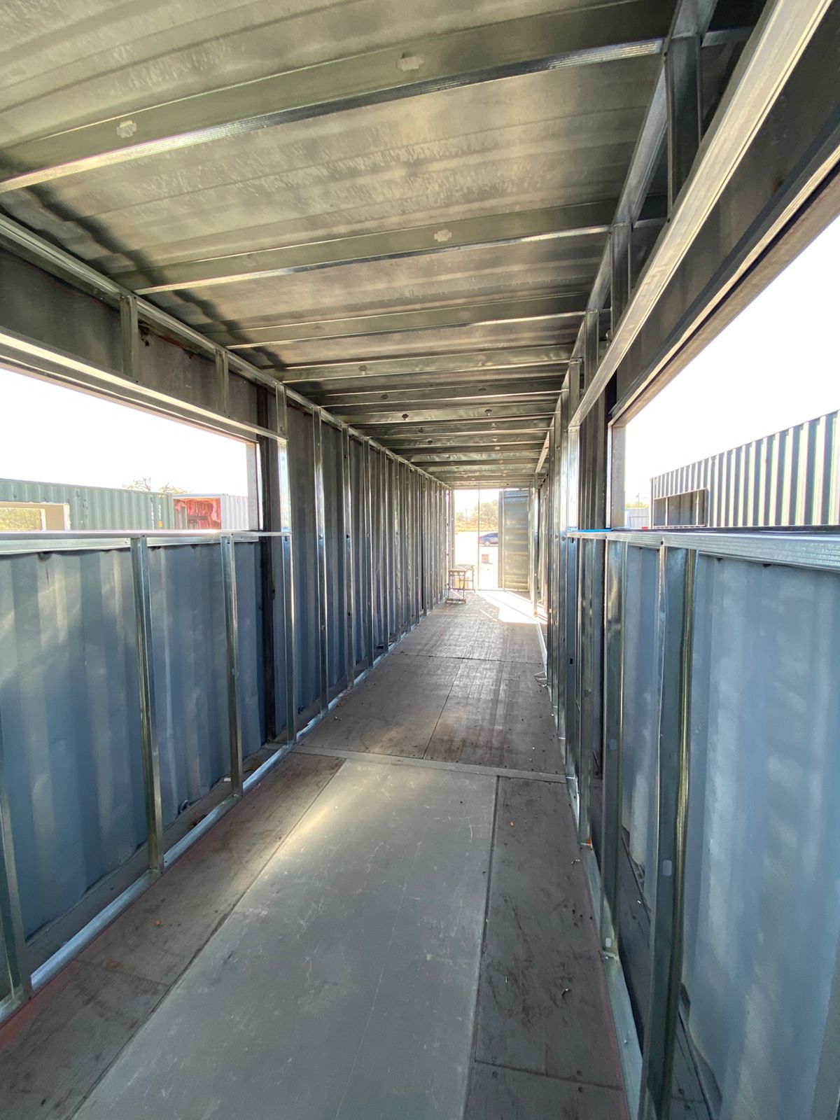 Vista longitudinal del contenedor con cortes laterales grandes ya ejecutados para ventanas corridas en ambos costados. Estructura de techo expuesta con vigas transversales y perfiles galvanizados perimetrales recién colocados.