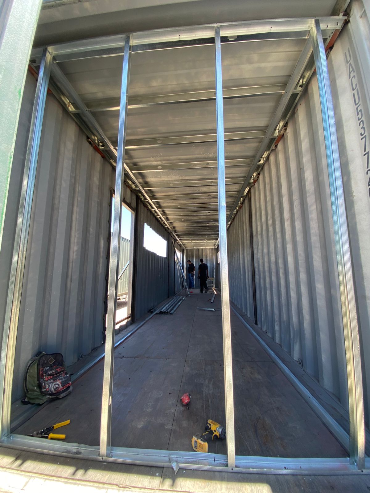 Interior de contenedor con perfiles galvanizados verticales montados como esqueleto previo a tablaroca, con vanos de ventana ya cortados a la mitad del muro. Se aprecian herramientas eléctricas (taladro, pinzas) y mochila de trabajo en el piso de madera original.