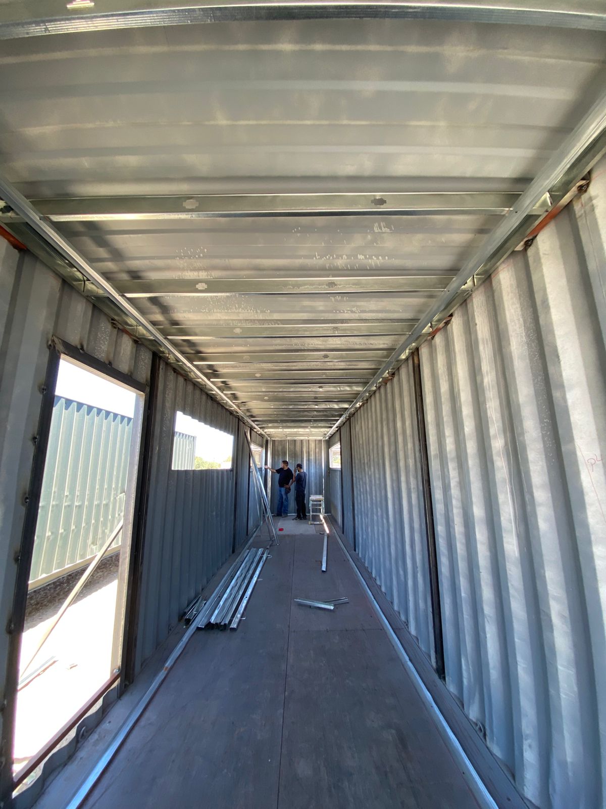 Vista longitudinal de contenedor con canaletas perimetrales de drywall ya fijadas en techo y piso, listas para recibir montantes verticales. Al fondo dos personas revisan plano o material acopiado en el pasillo.