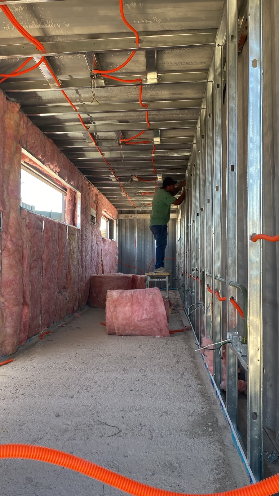 Trabajador sobre banco instalando aislante de fiberglass rosado entre perfiles galvanizados del muro lateral. Se aprecia el conduit naranja del cableado eléctrico ya tendido en plafón y la apertura de ventana sin marco. Etapa de envolvente térmica activa.
