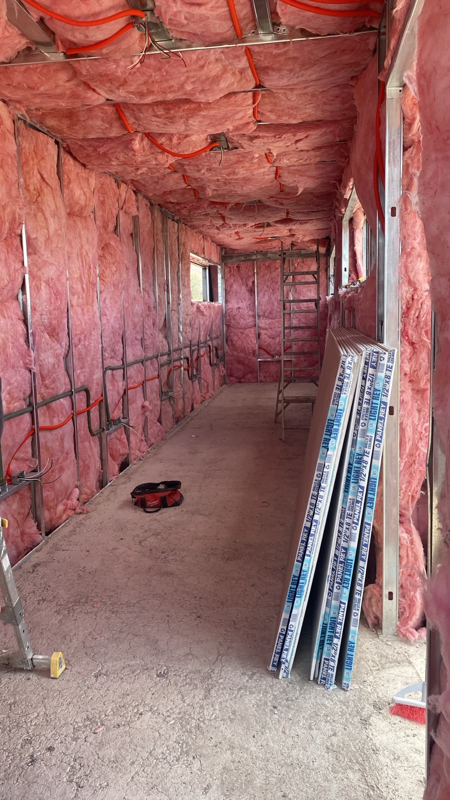 Interior del contenedor con aislante de fiberglass rosado R-18 instalado entre perfiles galvanizados en muros y techo. Se ve tubería conduit naranja para instalaciones eléctricas y paneles de tablaroca apilados listos para montar.