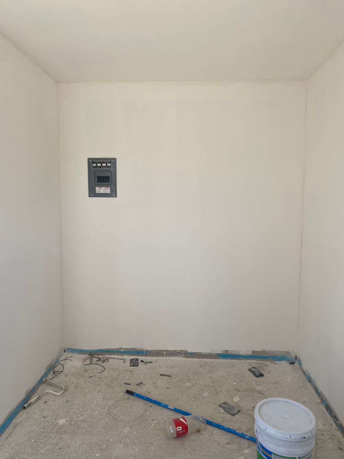 Cuarto pequeño con drywall ya pintado en blanco y centro de carga eléctrico montado en muro. En piso aún a nivel de firme se ven puntas de cable, herramienta de pintor y cubeta, antes del piso final.