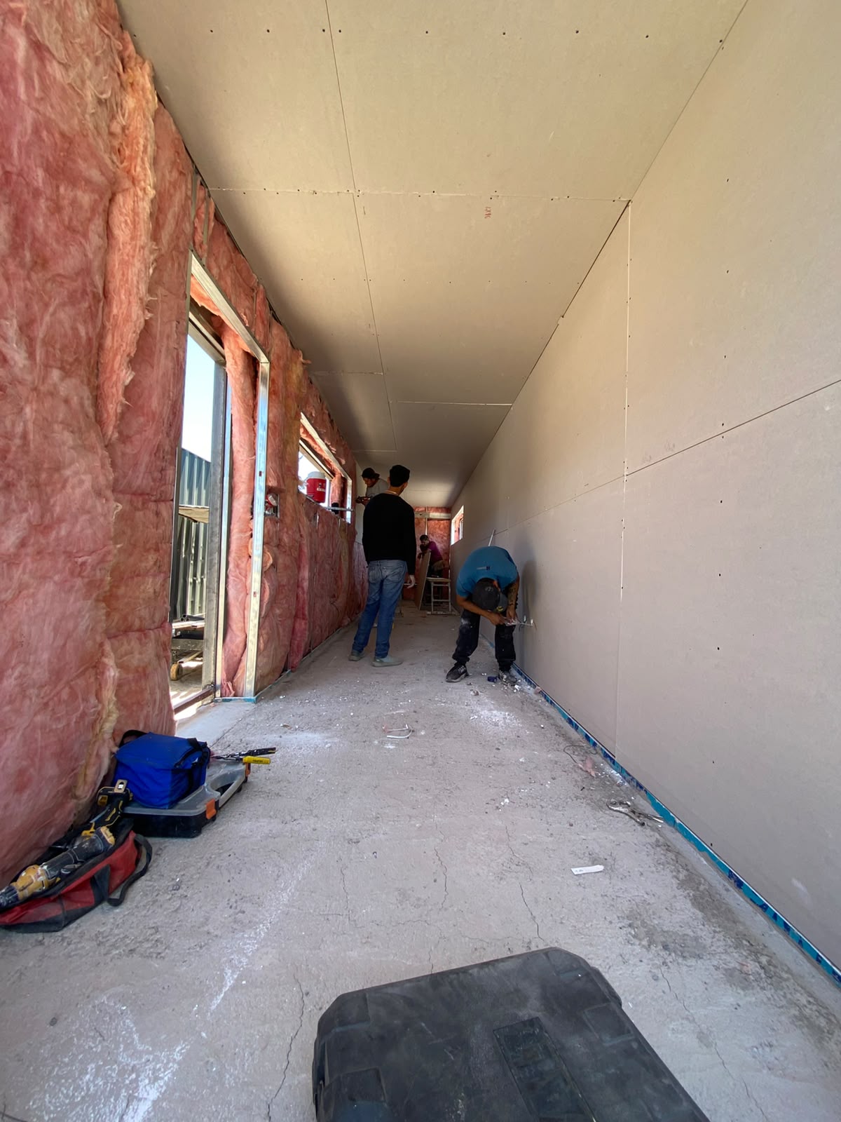 Pasillo interior con tablaroca ya instalada en muro derecho y plafón, contrastando con el muro izquierdo aún expuesto con fiberglass rosado. Dos trabajadores atornillando paneles cerca de la apertura de ventana al fondo.