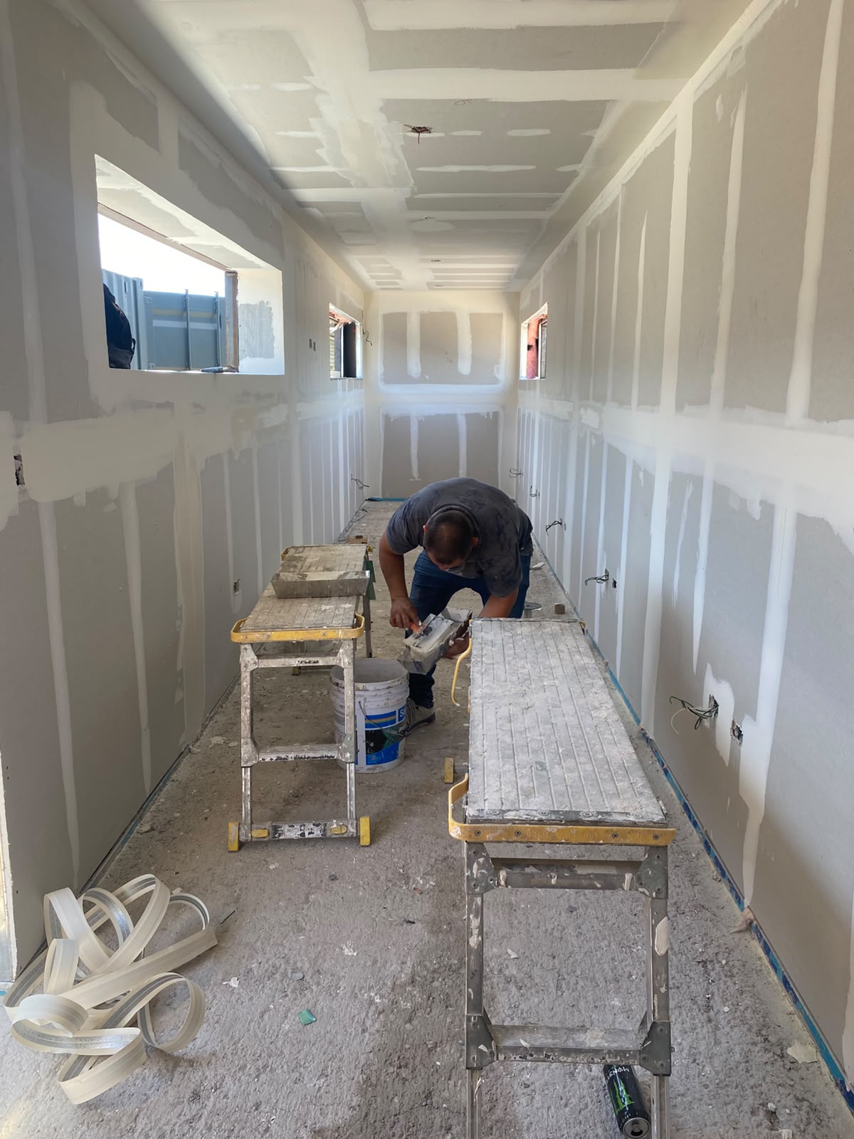 Interior de contenedor con tablaroca instalada y juntas resanadas, trabajador cortando perfiles sobre andamios improvisados. Las aperturas de ventana ya están recortadas y se ven puntos eléctricos pre-cableados saliendo de los muros.