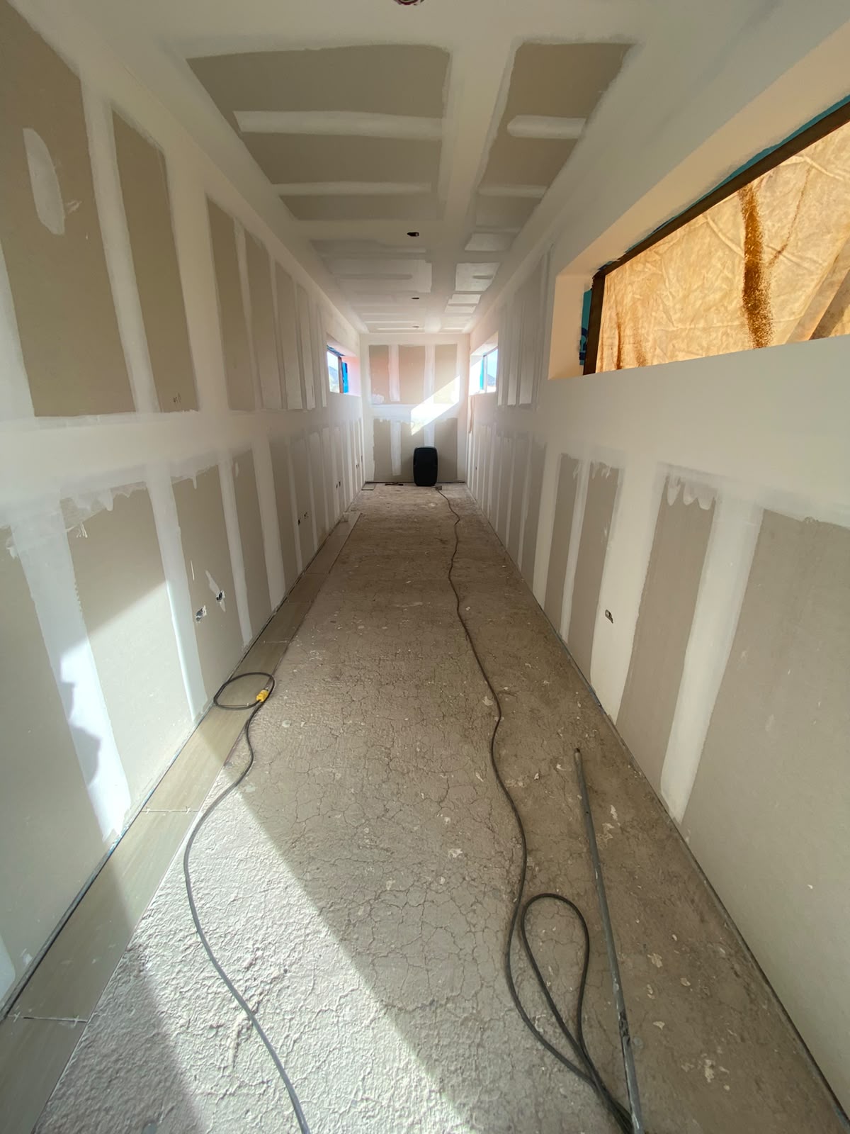 Interior longitudinal del contenedor con tablaroca instalada y juntas con compuesto aplicado sin lijar todavía, previo a pintura. Vanos de ventana cubiertos con tela kraft y cables sueltos en piso de concreto.