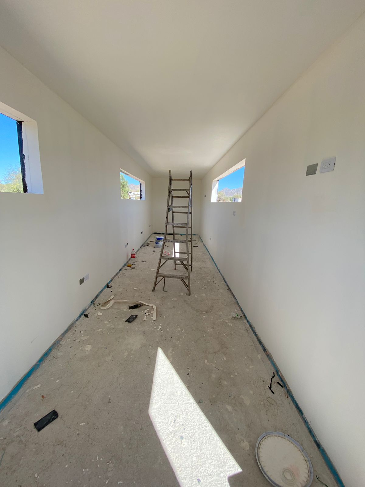 Interior del contenedor con drywall ya montado y pintado en blanco, tres ventanas abiertas hacia el cerro, contactos eléctricos colocados y piso aún en obra negra. Etapa muy avanzada salvo piso y carpintería.