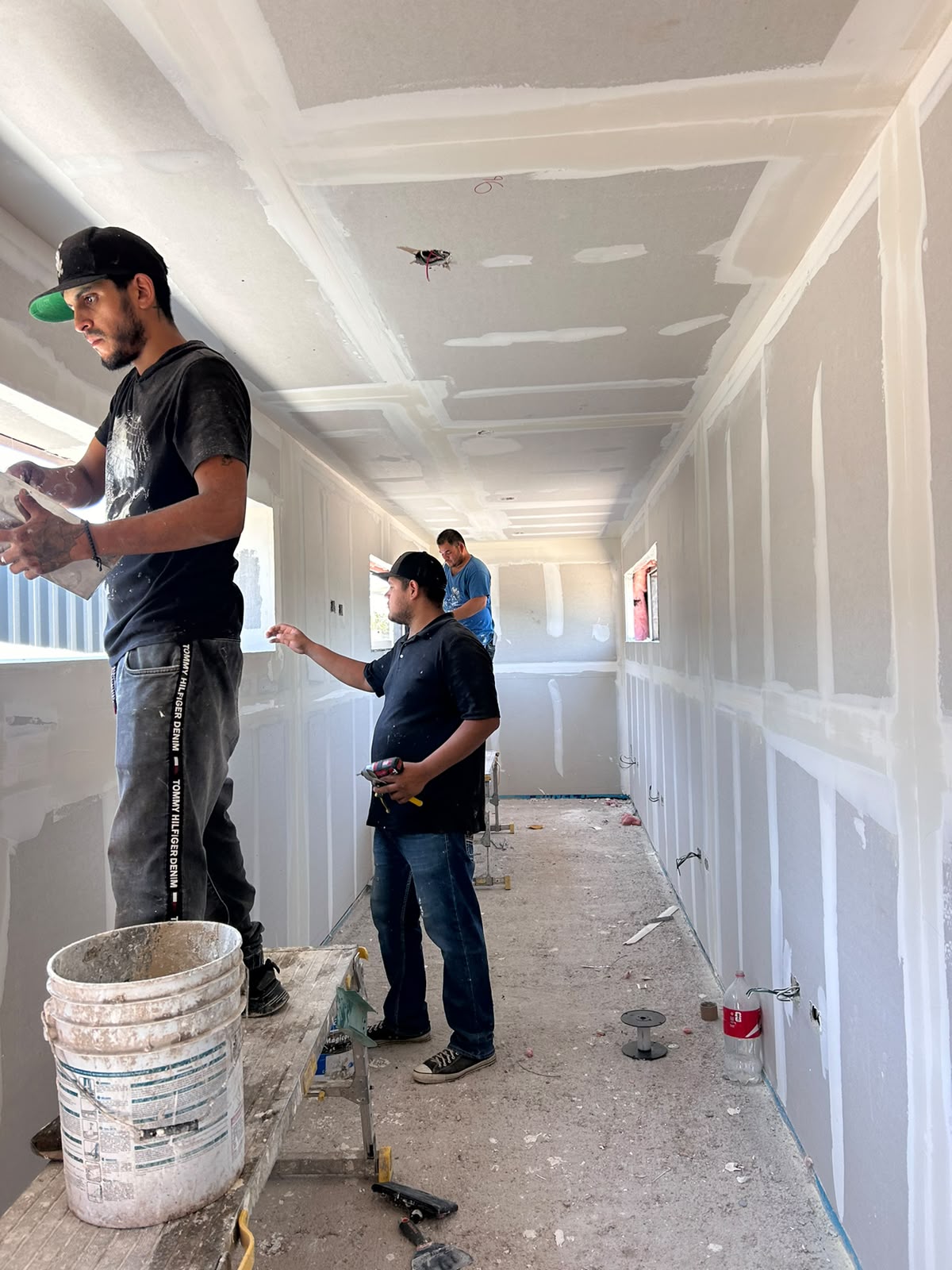 Tres trabajadores aplicando pasta de junteo y resanado sobre las uniones de tablaroca en muros y plafón, con cubetas de mezcla en piso. Etapa de acabado previo a pintura, con cortes de ventana abiertos hacia el exterior.