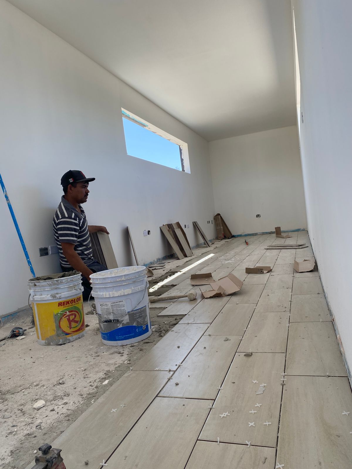 Módulo interior ya con drywall pintado en blanco y ventana horizontal alta. Trabajador asentando porcelanato imitación madera en proceso, con cubetas de adhesivo y crucetas niveladoras visibles.