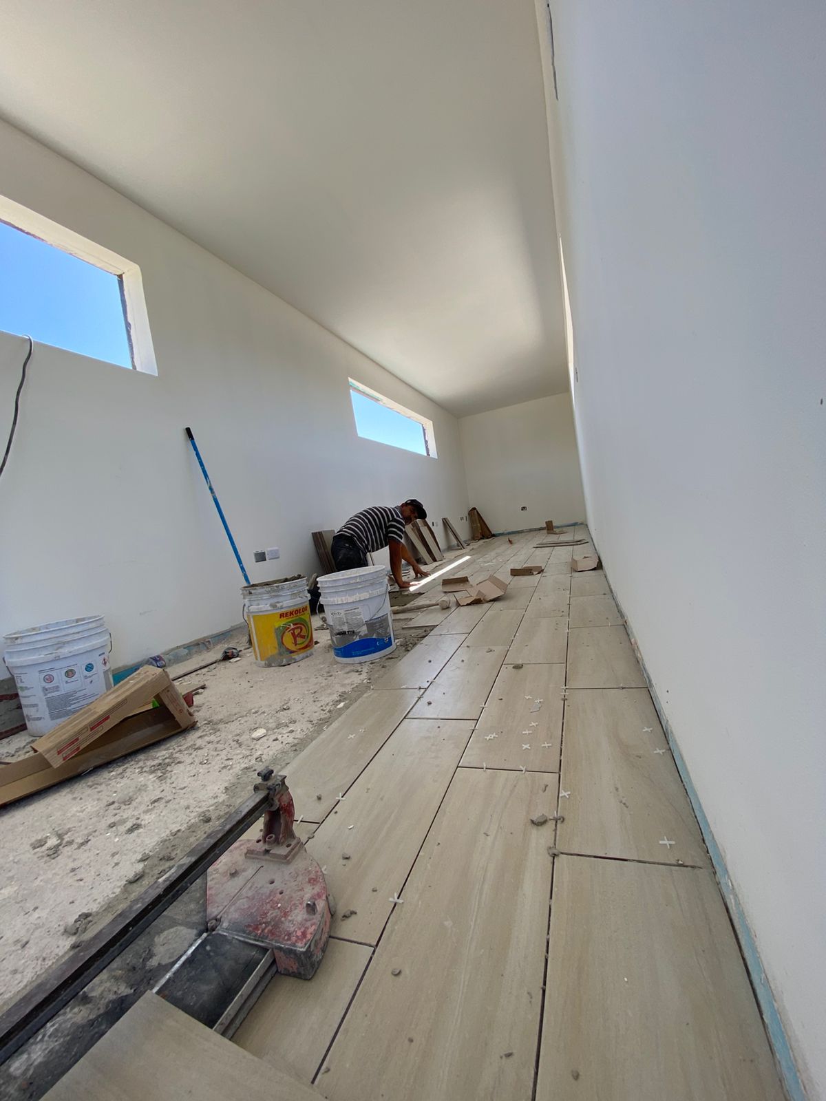 Interior alargado del contenedor en plena colocación de piso porcelanato imitación madera con crucetas separadoras visibles y un albañil cortando piezas al fondo. Muros ya pintados de blanco y dos ventanales horizontales altos dejan entrar luz natural.