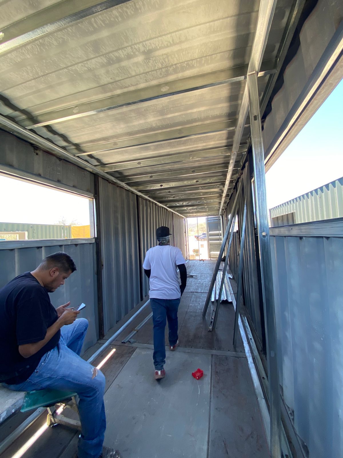 Dos trabajadores en faena dentro del contenedor en etapa de estructura: uno sentado revisando notas en primer plano y otro caminando hacia el fondo del módulo. Se ve un vano grande recién cortado a la izquierda.